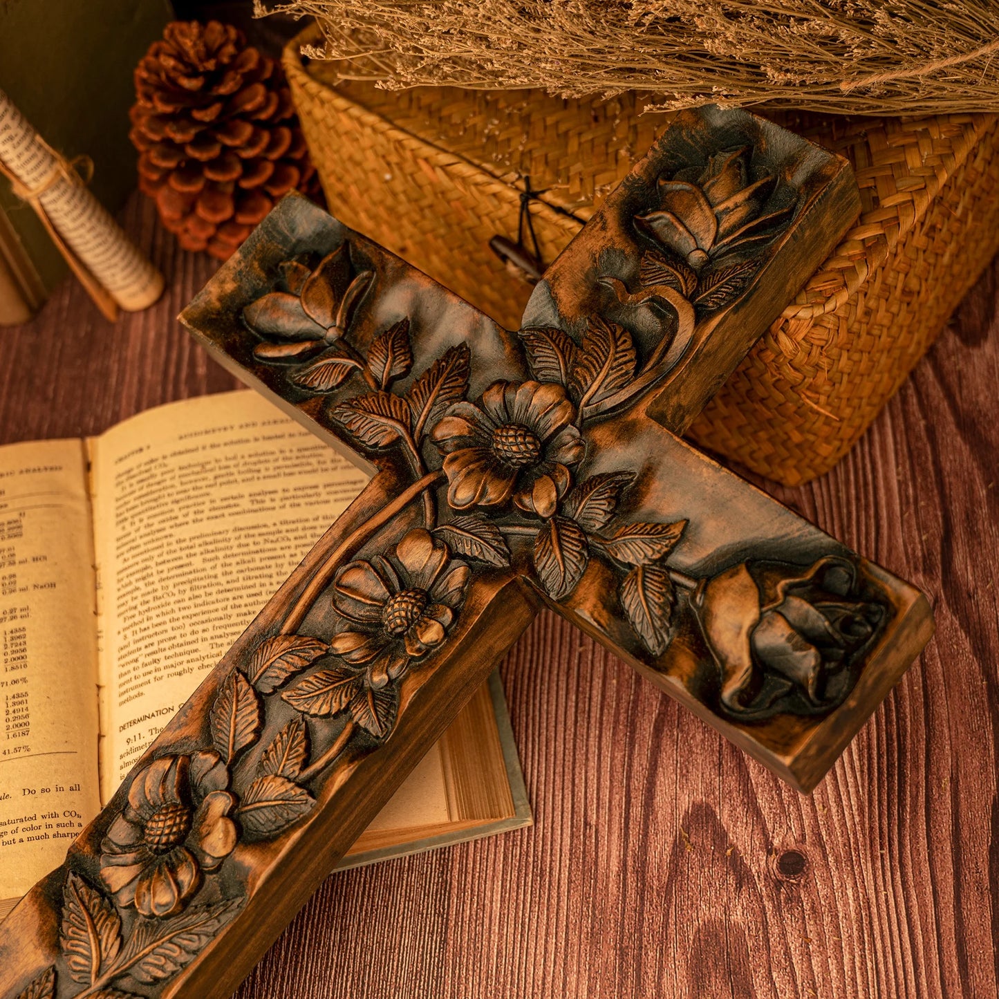 An overhead, close-up view of a large, dark-stained wooden cross with deep, detailed carvings of flowers and roses. The cross is placed on an open book, surrounded by rustic props including a wicker basket, pine cones, and rolled parchment on a dark wood table.