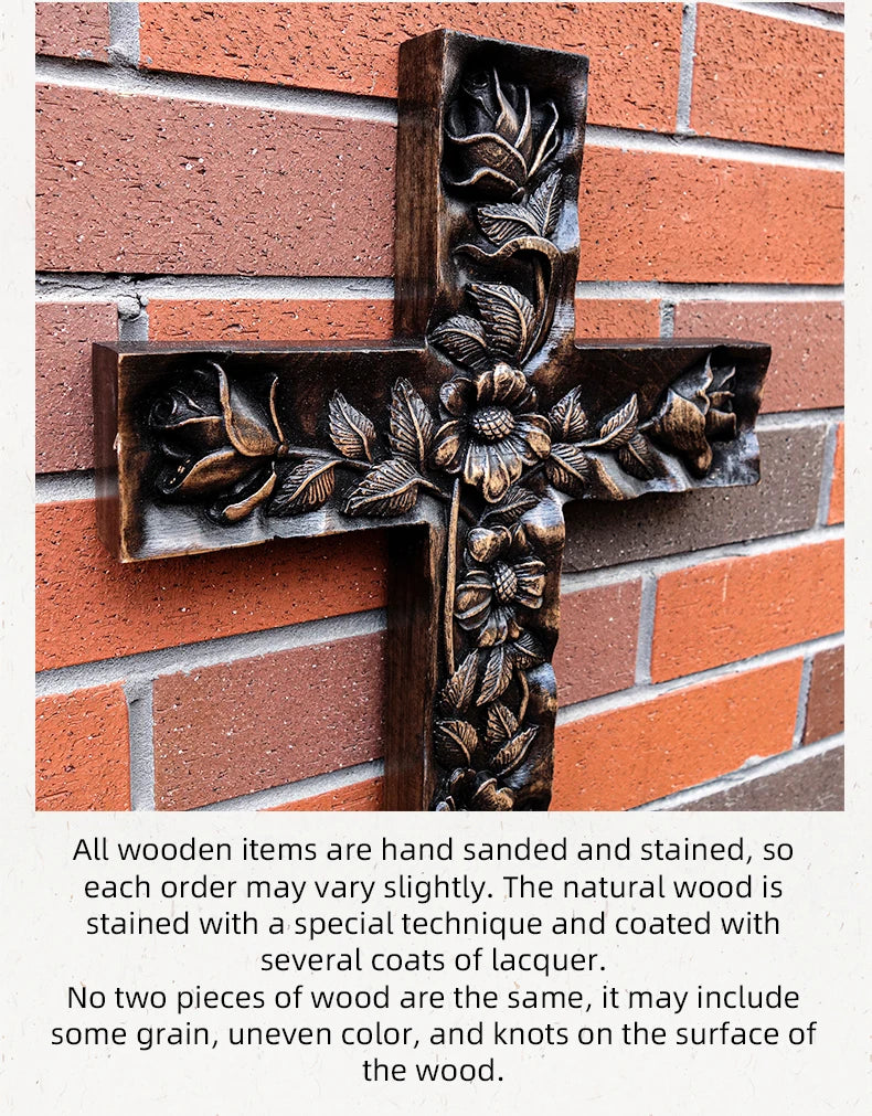Angled close-up of the dark wooden cross with deep floral and rose carvings, leaning against a red brick wall. The caption below describes the product as handmade, hand sanded and stained with lacquer, noting that each order may vary slightly due to the natural characteristics of the wood.