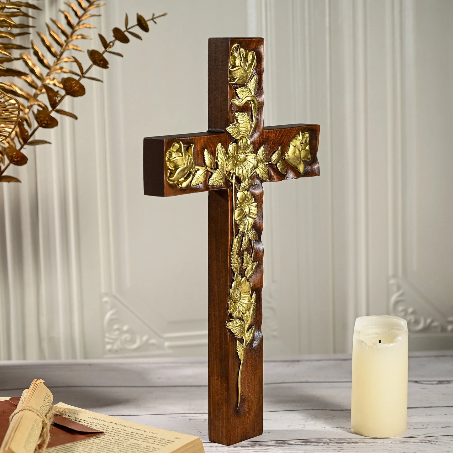 Side view of a thick, dark mahogany wooden cross featuring intricate gold floral and rose relief. The cross stands upright on a surface with a tall white pillar candle and gold botanical accents visible in the soft-lit background.