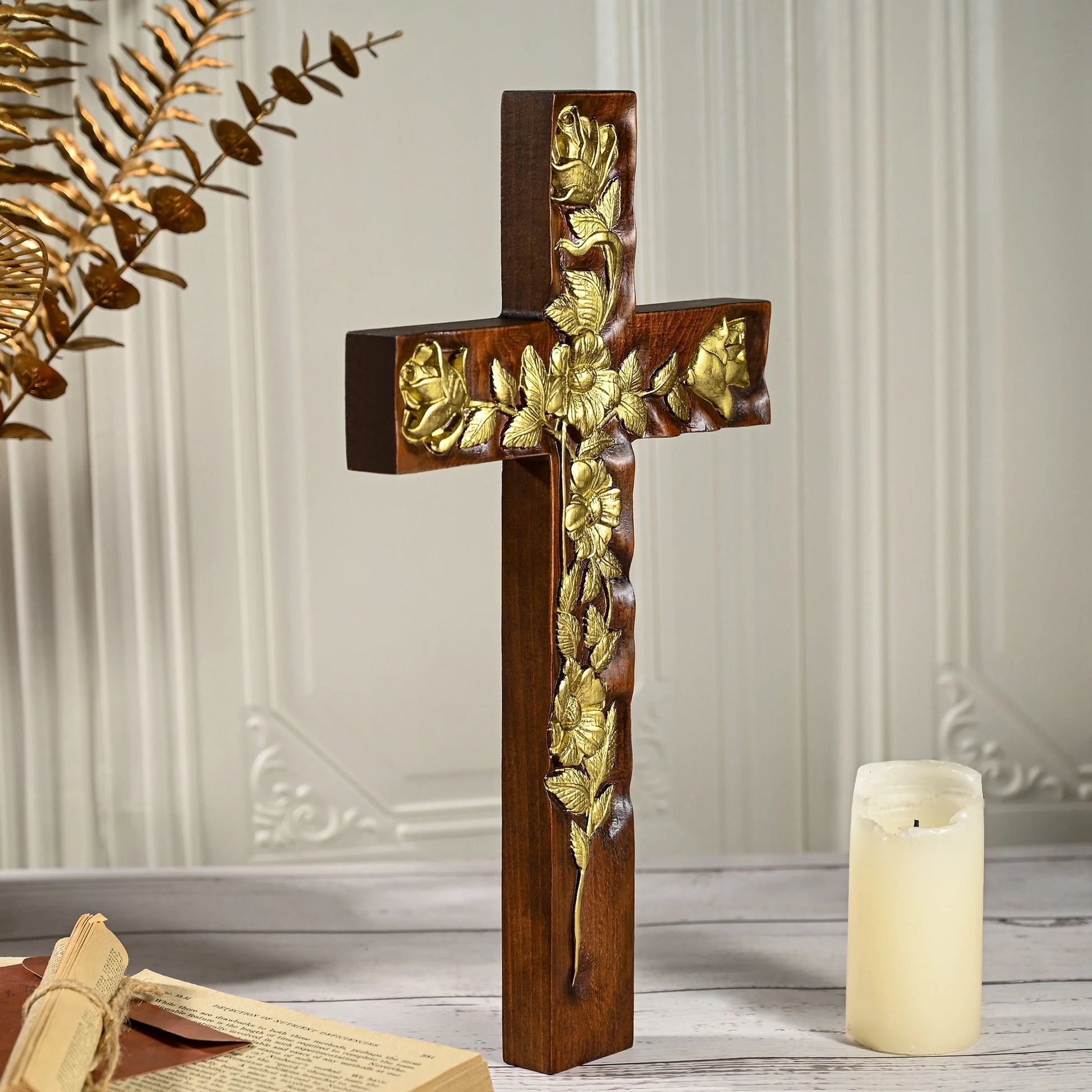 Side view of a thick, dark mahogany wooden cross featuring intricate gold floral and rose relief. The cross stands upright on a surface with a tall white pillar candle and gold botanical accents visible in the soft-lit background.