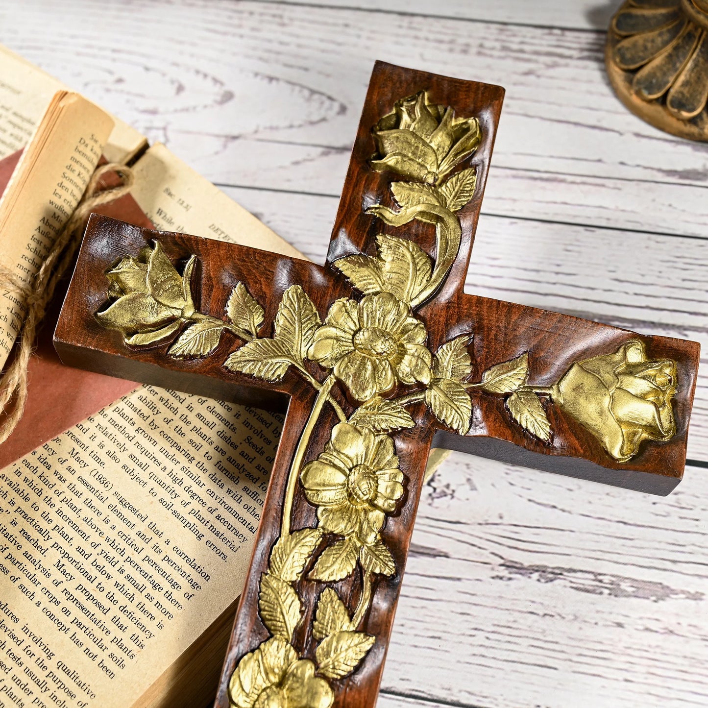 A close-up, overhead macro shot of a dark wooden cross featuring intricate, textured gold floral and rose relief. The cross is resting on a vintage open book with visible text and a rustic wood table surface, emphasizing craftsmanship and detail.