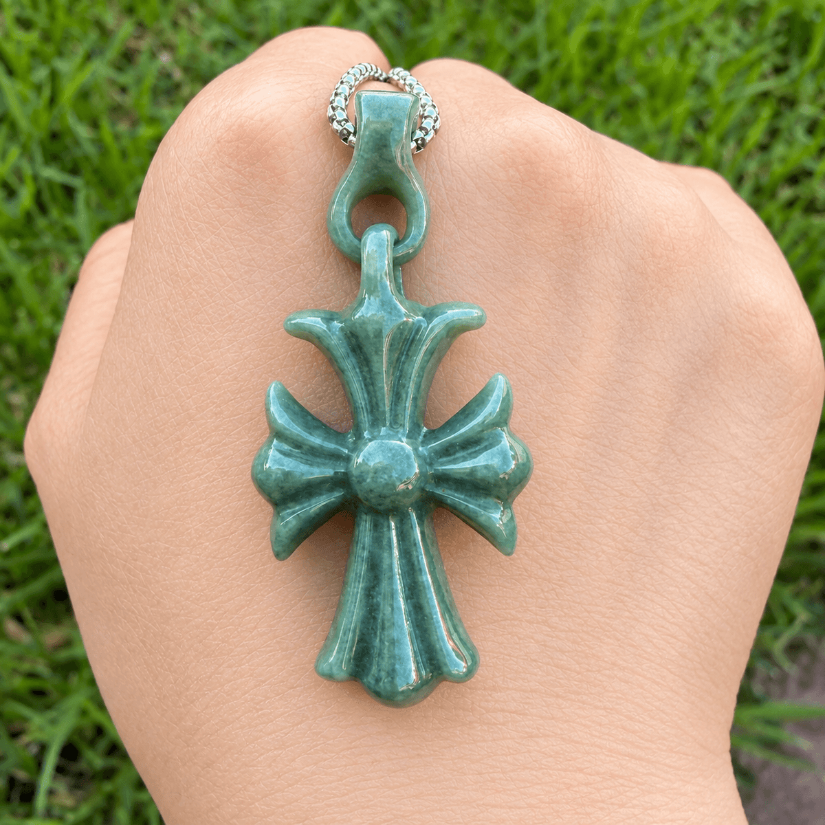 A carved cross pendant with a glossy teal-green finish, shown resting on a person's hand with a blurred green grass background.