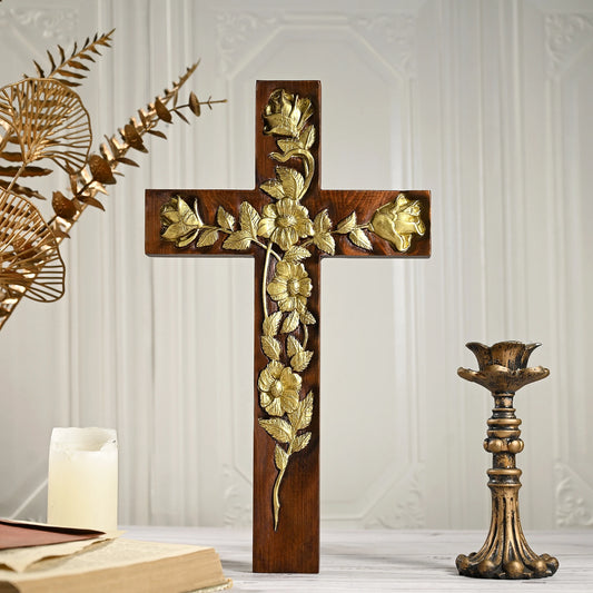 A freestanding dark wooden cross decorated with an elaborate gold floral and vine relief, set against a soft white panel background. A burning white candle and an open book are visible on a table surface in the lower left, creating a serene and devotional atmosphere.