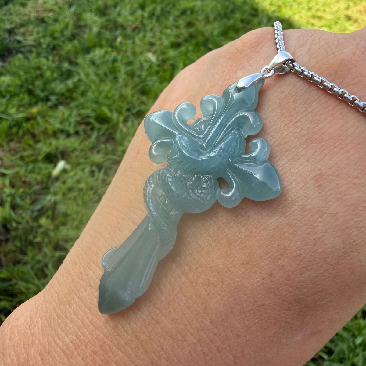 A close-up of a hand wearing a silver chain necklace with a carved light blue jade pendant, set against a blurred green grass background.