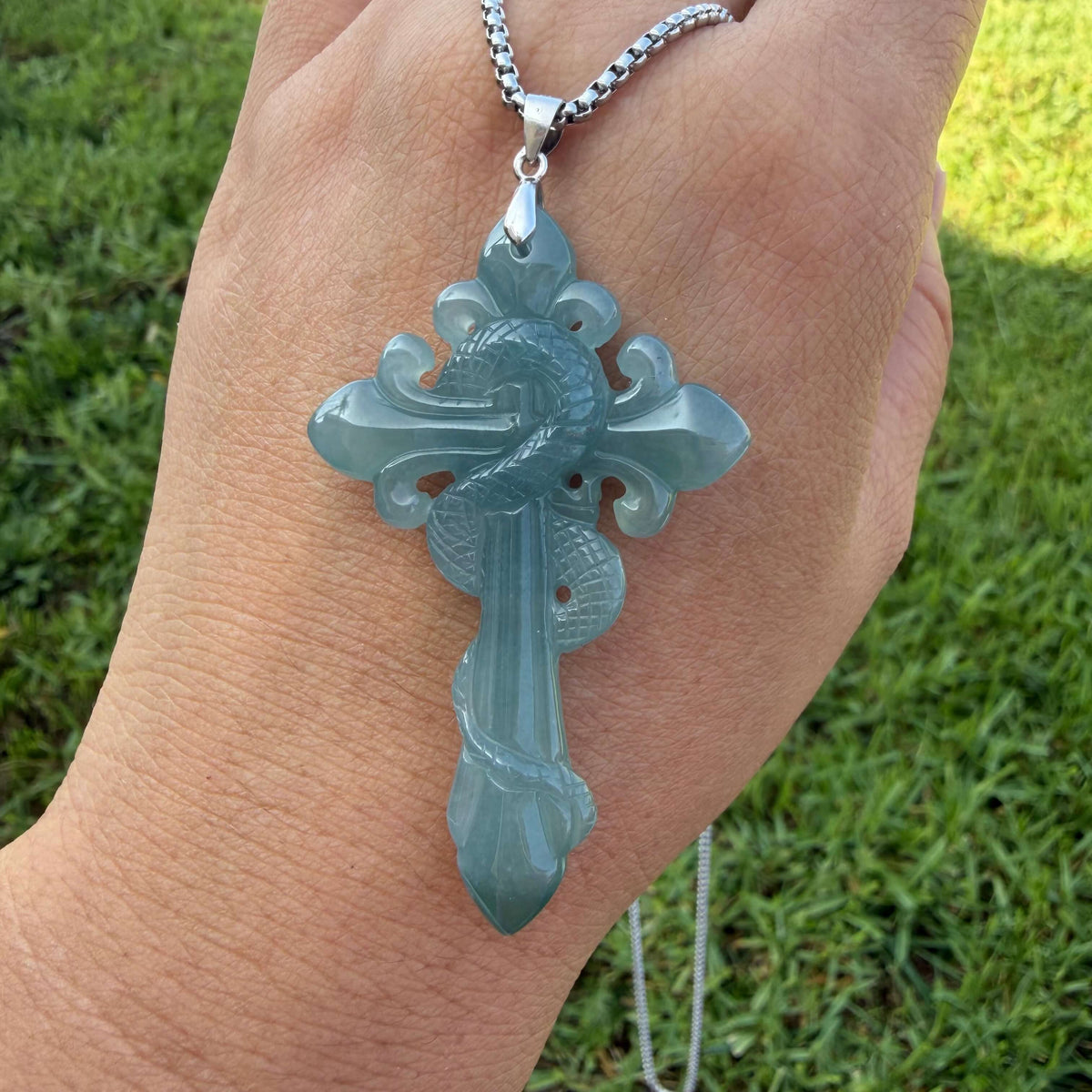 A carved cross pendant made of Blue water jadeite, hanging from a silver chain and worn on a person's hand, with a blurred green grass background.