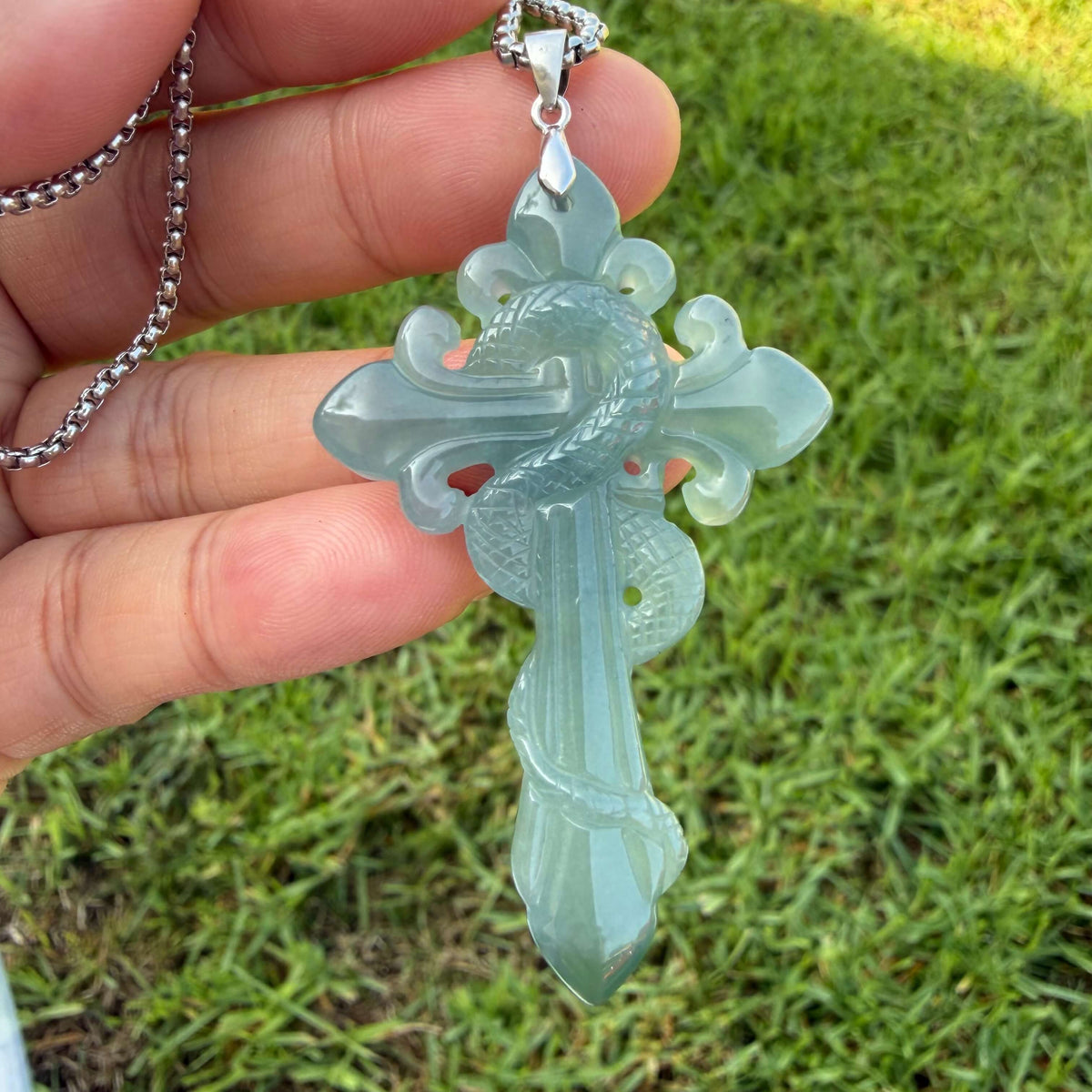 A hand holding a silver chain necklace with a carved light green cross Blue water jadeite pendant featuring a coiled dragon design, set against a blurred green grass background.