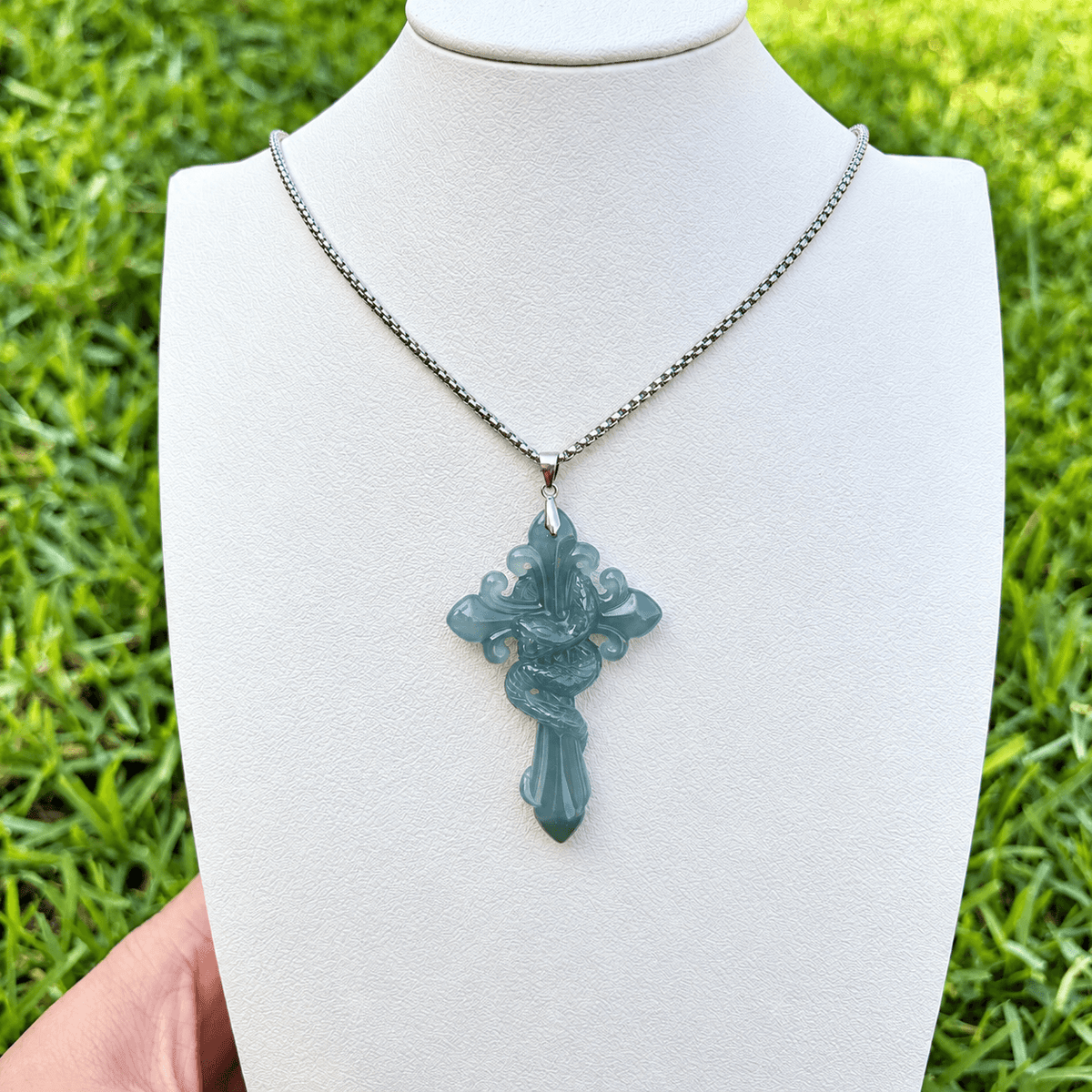 A silver chain necklace with a carved cross pendant made of a typa A jadetie, displayed on a white jewelry bust with a hand holding the left side and a green grass background.