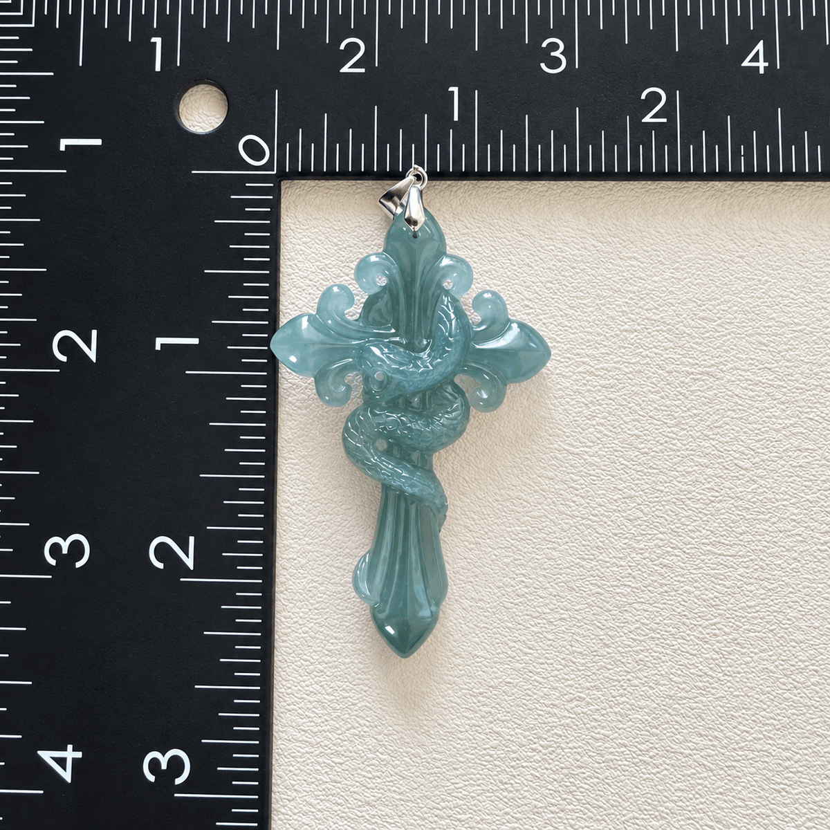 A carved cross-shaped pendant in light blue jadeite jade with a coiled snake wrapping through it, shown next to a black ruler for scale on a textured off-white background.