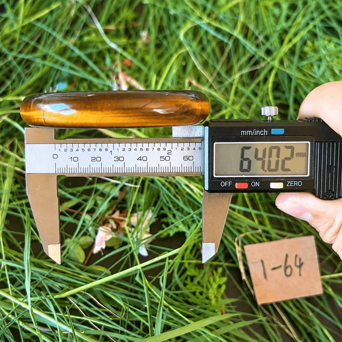 A hand holds a digital caliper measuring an amber-brown polished stone or resin sample placed on green grass. The caliper reads 64.