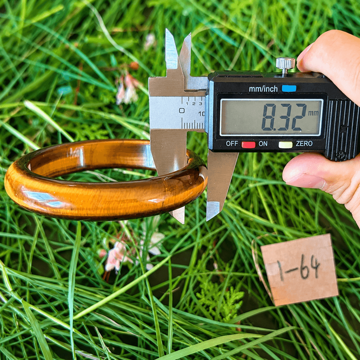 A hand holds a digital caliper measuring the width of a glossy brown wooden ring or bangle outdoors on green grass, with a small paper tag marked '1-64' nearby.