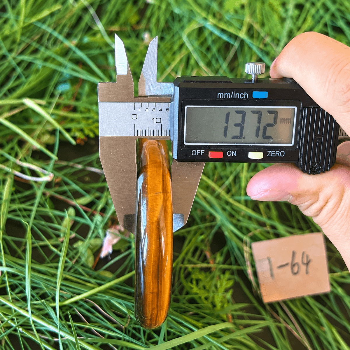 A hand holds a digital caliper measuring the width of a polished wooden paddle or handle, with green grass and water in the background and a small paper tag marked '1-64' nearby.