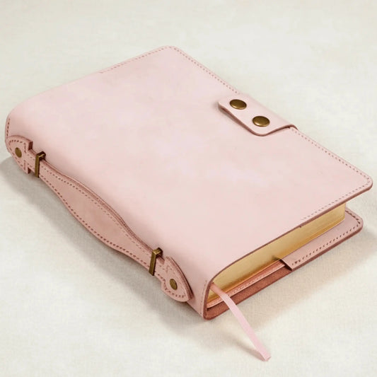 A close-up photo of a Bible encased in a light pink leather cover. The cover is smooth and features an adjustable side leather strap with brass hardware, and a front flap secured by two brass snap buttons. The Bible's page edges are gilded (gold-colored), and a pink ribbon bookmark is visible.