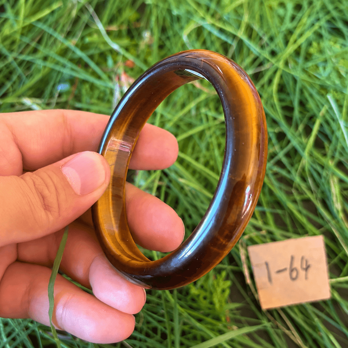 A hand holds a polished tiger's eye bangle against a background of green grass, with a small wooden tag marked '1-64' nearby.