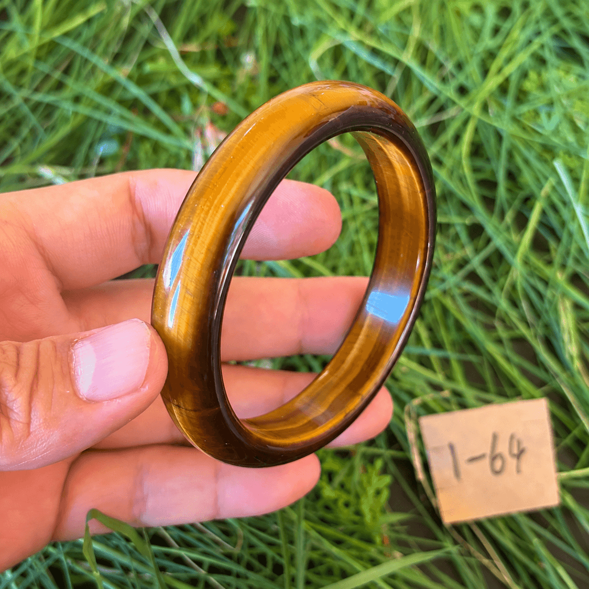 A hand holds a polished tiger's eye bangle ring against a background of green grass, with a small wooden tag marked '1-64' nearby.