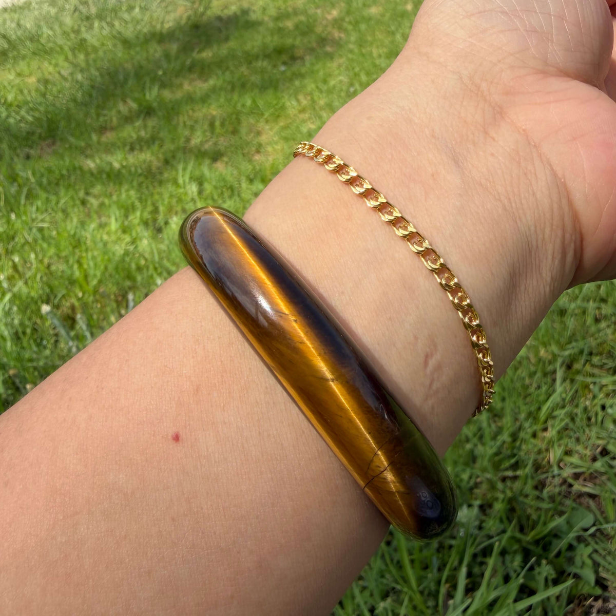 A close-up of a person's wrist wearing a polished tiger's eye bangle and a gold chain bracelet, set against a blurred green grass background.