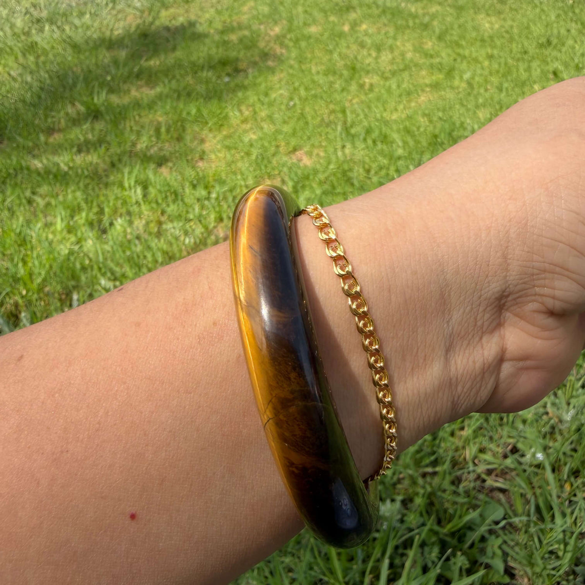 A person's wrist wearing a large, polished tiger's eye bangle alongside a delicate gold chain bracelet, set against a blurred green grass background.