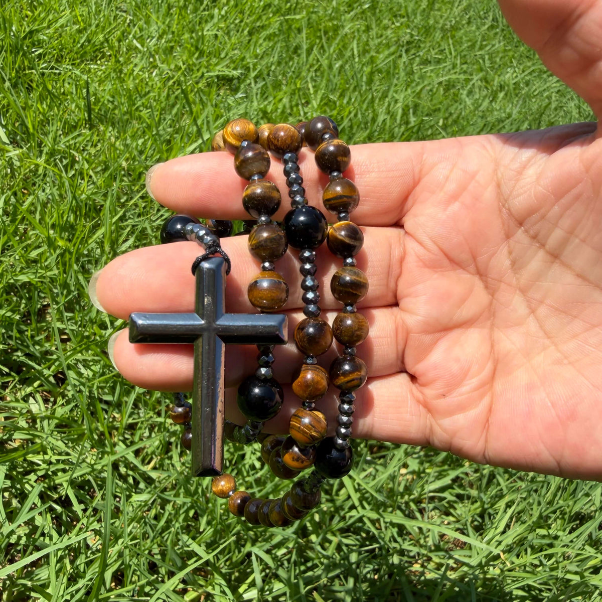 A close-up of a hand holding a rosary with dark metallic cross and beads, including brown and black tiger's eye beads, against a background of green grass.